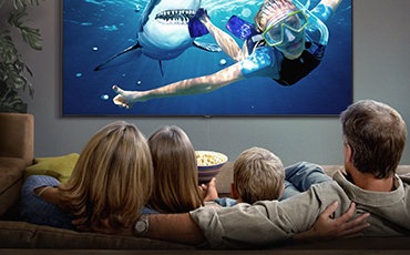 Es sind 2 Bilder zu sehen. Auf dem linken Bild schauen 4 Personen einen spektakulären Film auf einem Super Big TV. Auf dem Bildschirm ist eine Frau beim Gerätetauchen zu sehen, die vom einem Hai verfolgt wird. Auf dem rechten Bild ist ein gemütliches graufarbiges Wohnzimmer aus Vogelperspektive zu sehen. Ein Mann sitzt auf einer Couch und sieht eine wunderschöne Stadt bei Nacht auf dem Super Big TV.