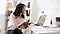 Une femme assise à un bureau tenant une tablette, regardant un écran d’ordinateur portable dans un appartement lumineux.