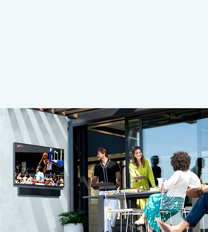 La nouvelle télévision Samsung The Terrace et la barre de son The Terrace sont visibles en diagonale sur un mur. Et les gens préparent le dîner dans le jardin.