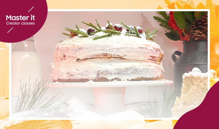 Un pastel de hermosas capas cubierto con glaseado blanco, bayas y agujas de pino, sobre un soporte para pasteles en una cocina. La frase Master it creator classes se asoma por la esquina superior izquierda.