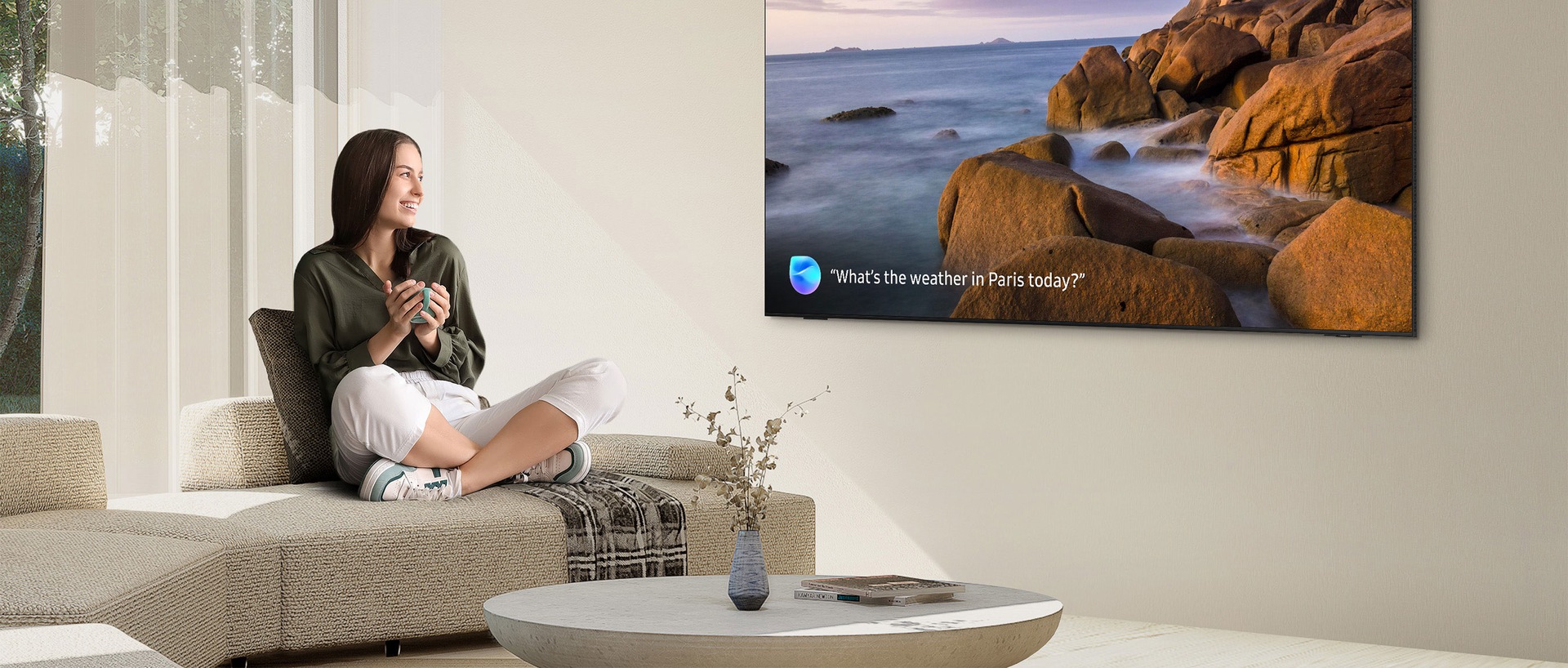 En una sala de estar, una mujer sonríe a un TV en la pared. La pantalla del TV muestra una escena al aire libre de acantilados y agua con el mensaje ¿Cuál es el clima en París hoy?