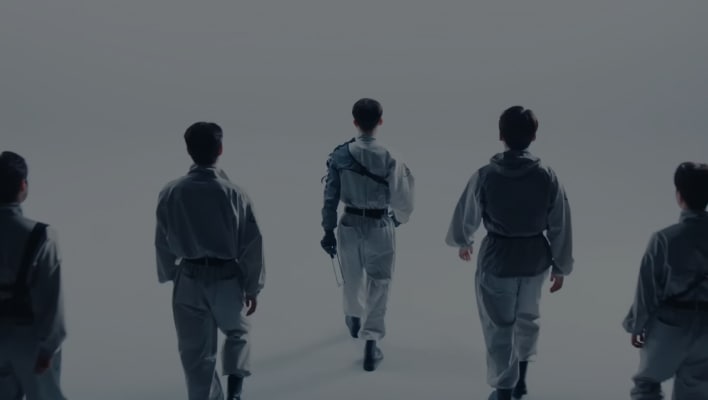 A group of 5 men walk away from the screen in a white room.