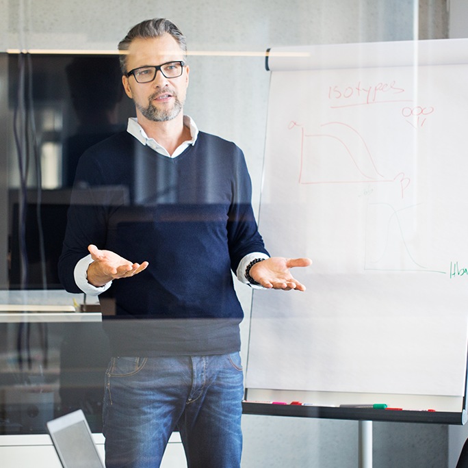 Ein Mann steht mit seitlich zu einem Flip-Chart mit einem aufgemalten Diagramm und spricht mit leicht nach vorne geöffneten Händen zu seinen Zuhörern außerhalb des Bilds