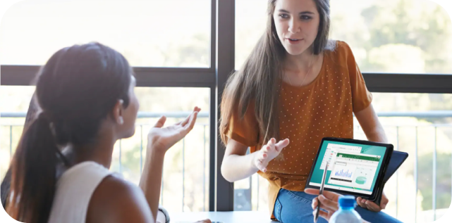 Zwei Damen sprechen miteinander. Eine sitzt mit dem Rücken zum Betrachter an einem Tisch, die andere sitzt halb seitlich vor ihr und hält ein Samsung Tablet mit dem Display nach vorne gerichtet in ihre Richtung.