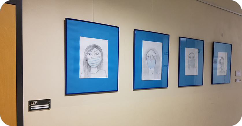 Eine Reihe von selbstgezeichneten Bildern von Menschen mit Atemschutzmaske hängen an einer Wand.