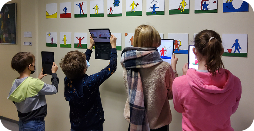 4 Kinder stehen mit Tablets in der Hand vor einer mit selbstgemalten Bildern behangenen Wand, auf die sie ihre Tablets richten.