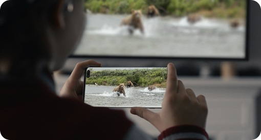 Ein junges Mädchen hält ein Galaxy Note10 mit beiden Händen. Auf ihrem Handy ist eine Szene mit Bären im Wasser zu sehen, die gleichzeitig auf dem Fernseher vor ihr läuft