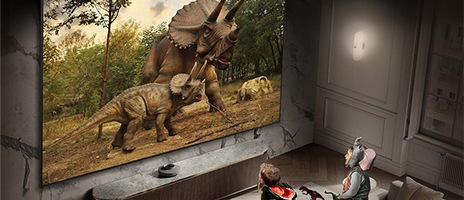 Zwei kleine Kinder in Tierkostümen beobachten vertieft zwei Dinosaurier auf dem großen Supersize-Display von Samsung im Wohnzimmer.