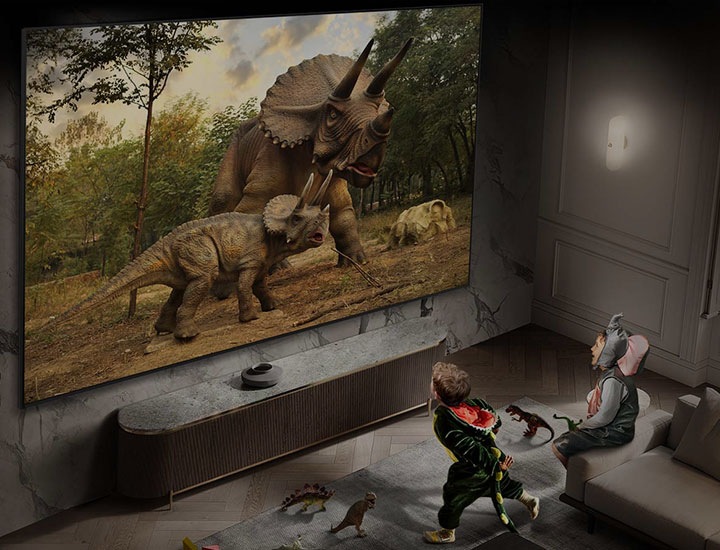 Zwei kleine Kinder in Tierkostümen beobachten vertieft zwei Dinosaurier auf dem großen Supersize-Display von Samsung im Wohnzimmer.