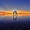 Et par står og laver sjove fagter mod hinanden på et reflekterende underlag med en farverig solnedgang i baggrunden