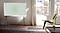 A Cloud White model of The Serif sits on a black stand in a bright and elegantly simple living room, with an area rug, a stylish three-legged green chair, and large windows. The image on screen is Leaf Ambient Mode.
