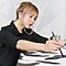 A light-haired woman in a black top is sitting and painting a large piece of art. She is holding a brush on her right hand and touching the canvas to paint with a black ink.