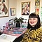 A short-haired woman with dark hair is sitting at her desk smiling wearing a leopard print top. On her desk are art materials and there are colourful art prints in the background.