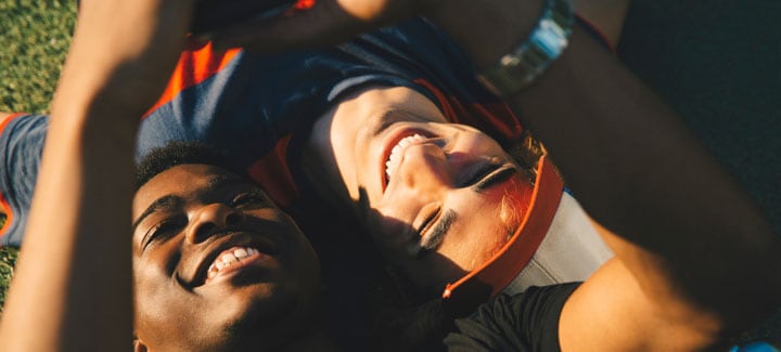 Dos amigos tumbados juntos en el suelo en direcciones opuestas, que están mirando a un smartphone que sostiene uno de los hombres, mientras el sol brilla sobre ellos.