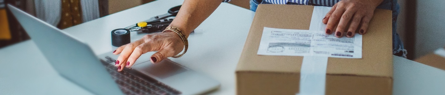 Un primer plano muestra a una mujer de pie ante una mesa. Su mano izquierda reposa sobre un paquete que tiene una etiqueta de correos y con su mano derecha está utilizando un ordenador portátil. Sobre la mesa también hay cinta adhesiva, tijeras y un cúter.