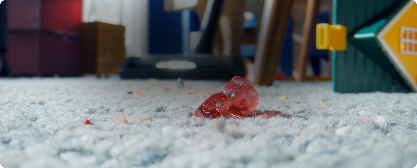 Una mujer utiliza el aspirador Samsung Jet para limpiar una alfombra gris en la habitación de un niño. En la alfombra hay una casa de juguete con tejado verde y un osito de goma rojo. Cuando la mujer mueve la aspiradora hacia el osito de goma, la escena cambia a un primer plano de la cara del osito de goma mientras es aspirado por la aspiradora junto con el polvo de la alfombra.