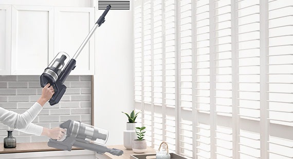 Una persona levanta la ligera Jet 65 con una mano para limpiar el hueco de ventilación alto con la herramienta para hendiduras extensible y limpia la encimera con la herramienta combinada.