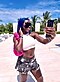 Une femme aux cheveux bleus portant un débardeur blanc et un short en jean pose pour un selfie.