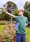 Un homme portant un t-shirt vert et un jean prend un selfie devant des fleurs dans une rue de quartier.