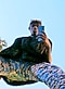 Un homme portant un pantalon bleu et jaune et une fourrure prend un selfie miroir vers le bas devant un ciel bleu.