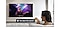 A woman sitting on a white couch in a brightly lit living room watching a concert video on a wall mounted Smart TV. The woman activates the voice assistant by saying "Turn down the volume" into the Smart TV remote she's holding.