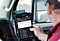 A man sitting in a vehicle pressing his thumb on a rugged black Samsung tablet that is held in a tablet holder