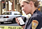 A female police officer standing at the crime scene talking with a black rugged Samsung smartphone in her hand.