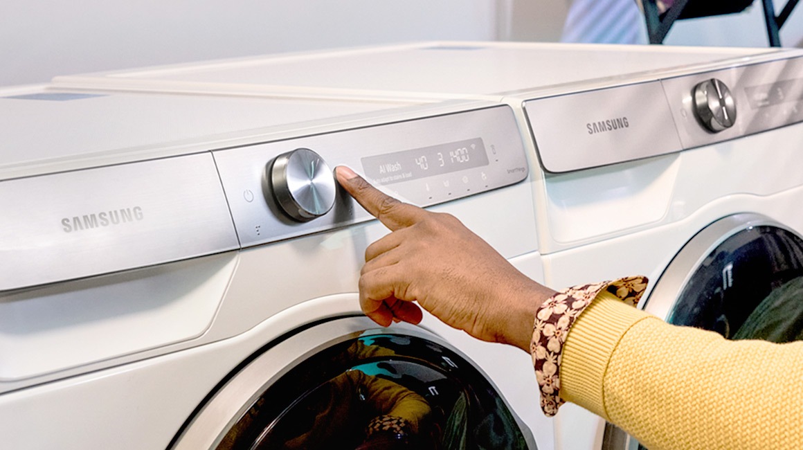 Ένα χέρι πατάει το κουμπί έναρξης του λευκού πλυντηρίου της Samsung.
