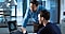 In a cool-tone modern office setting, two men are looking at a Galaxy Book screen and discussing its contents. One man is sitting at a desk and pointing at the screen, while the other is leaning over the desk to see the screen.