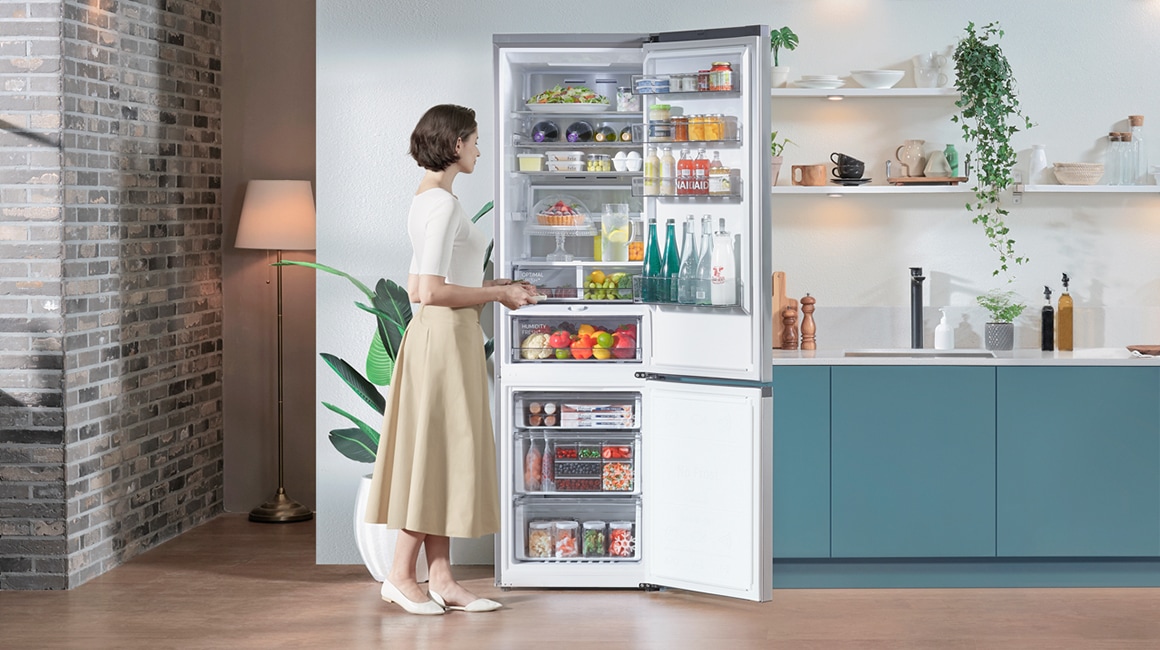 Egy modern, minimalista konyhában egy kadettkék konyhaszekrény mellett, egy nő éppen kinyit egy Samsung hűtőszekrényt, aminek a nagy hűtőrészében rengeteg étel van szépen elrendezve.