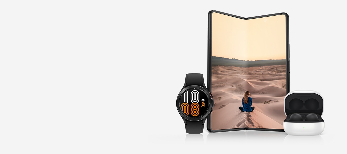 Galaxy Watch4 with an orange and white watchface. Galaxy Z Fold3 5G unfolded and seen from the Main Screen with a photo of a woman sitting in the middle of sand dunes on screen. Galaxy Buds2 seen with the cradle open and the earbuds in place.