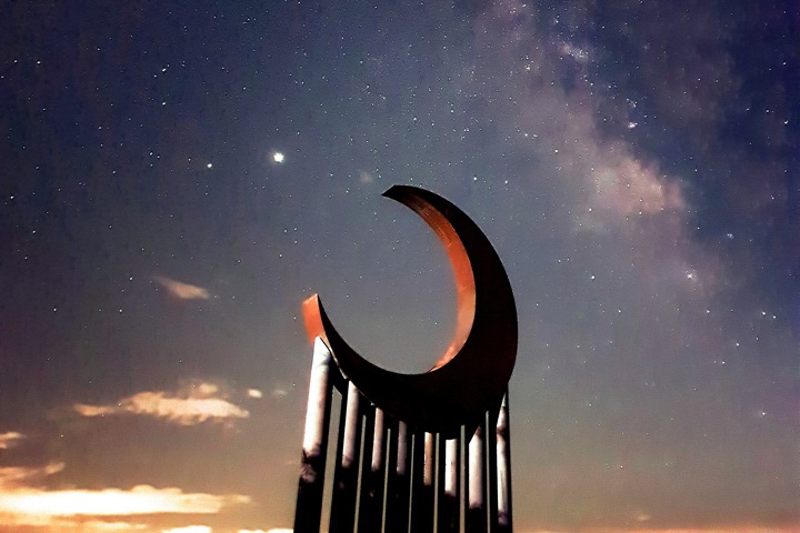 Sculpture of a moon, supported by columns against a starry night sky during sunset
