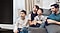 A laughing and smiling family of four are sitting closely together on a sofa enjoying a big screen TV.