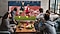 A group of five men sit around a coffee table while watching and cheering excitedly for a soccer game on a Super Big TV.