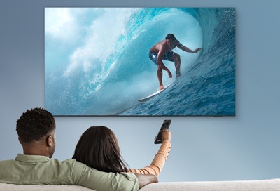 A couple on a couch watching a surfer on a large wave displayed on their Samsung Smart TV, using a remote to navigate.