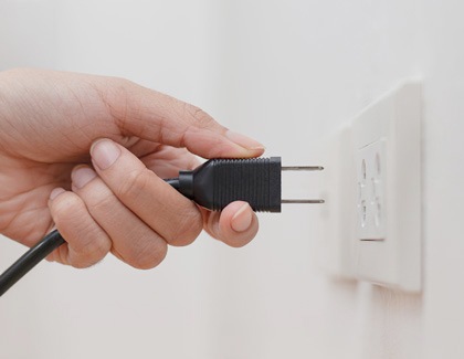 A person's hand holding an unplugged electrical plug near a wall socket.