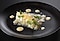 At the center of a round black plate, strips of potato are layered on top of cod that has been carefully mashed. To complete the elegant plating, a white sauce has been dripped on top of the dish and drizzled artfully around the plate, all accentuated by a sprinkling of chervil.
