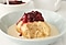 There is a dumpling in a white plate, and roasted plum compote on its top. And vanilla custard drizzled around the dumpling.