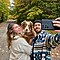 A bearded man in a colourful blue coat takes a family selfie with Galaxy S21 Ultra 5G. The selfie also featuring his brown dog and a young woman in a white knit sweater on a peaceful forest road bordered by beautiful autumn foliage.