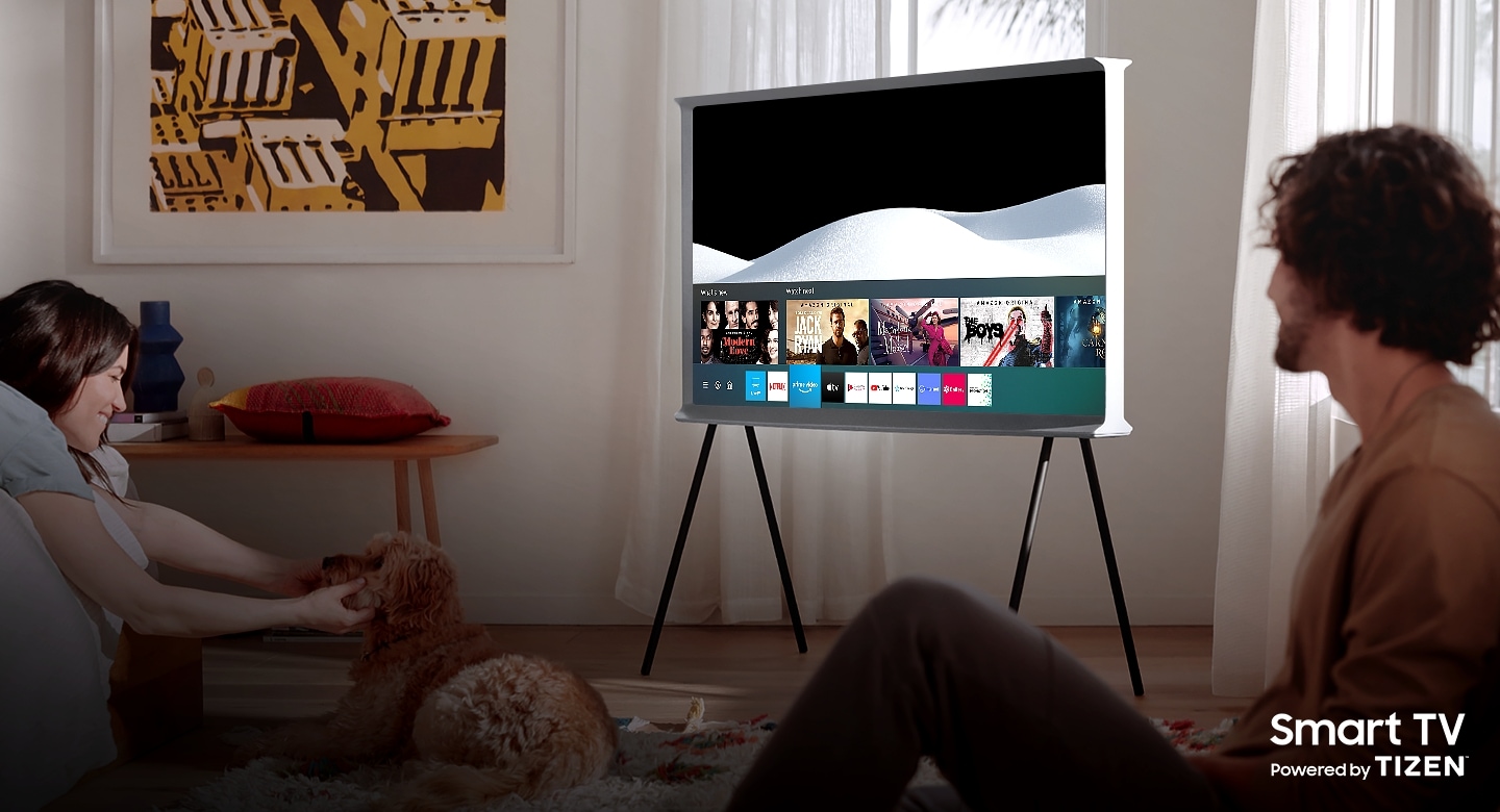 A man and a woman are enjoying their time together in a living room.  A Cloud White The Serif sits in the center of the room with the screen Samsung Smart Hub open.The Serif TV is Smart TV powered by Tizen.