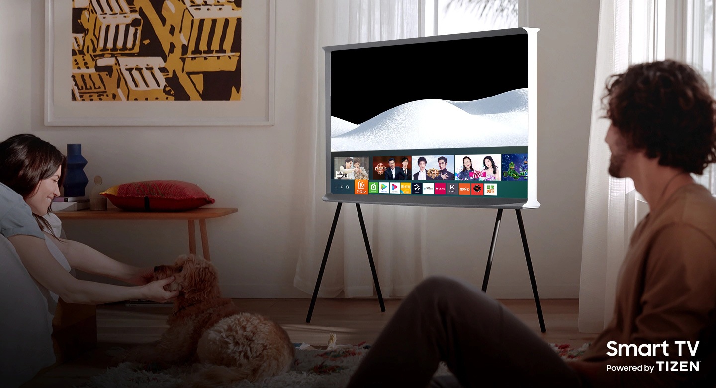 A man and a woman are enjoying their time together in a living room.  A Cloud White The Serif sits in the center of the room with the screen Samsung Smart Hub open.The Serif TV is Smart TV powered by Tizen.