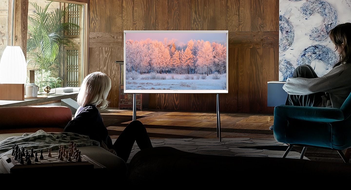 Two women look at a Cloud White model of The Serif TV on a stand in a stylish living room. The image on The Serif’s screen shows a winter scene with a forest and snow. It zooms in to focus The Serif