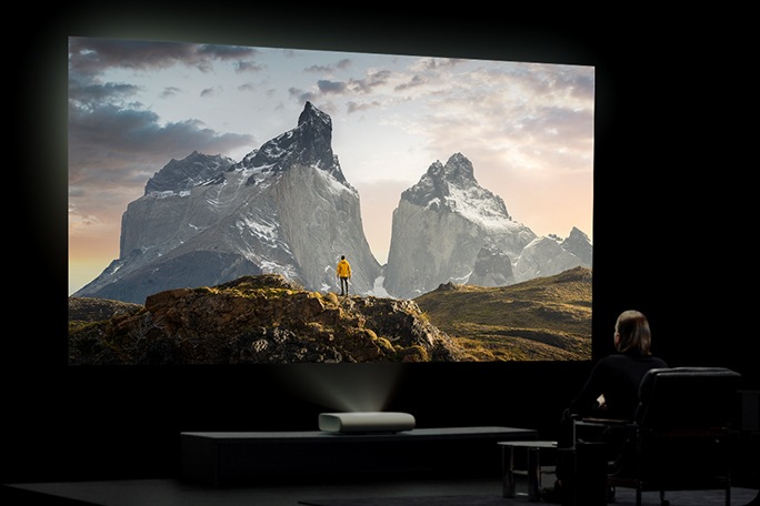 شخصی در یک اتاق تاریک و شبیه سینمای خانگی، صحنهای از منظره را روی یک صفحه نمایش بزرگ که توسط The Premiere 9 نمایش داده میشود، تماشا میکند.