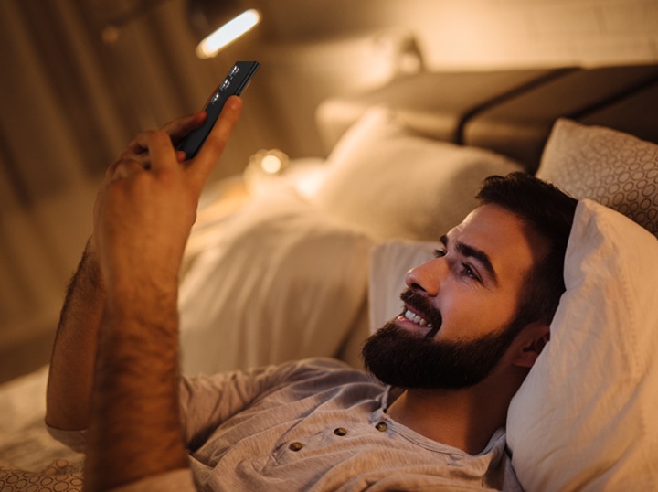 رجل يستخدم هاتفه الخلوي أثناء استلقائه على سريره مع أضواء أرضية مضاءة.