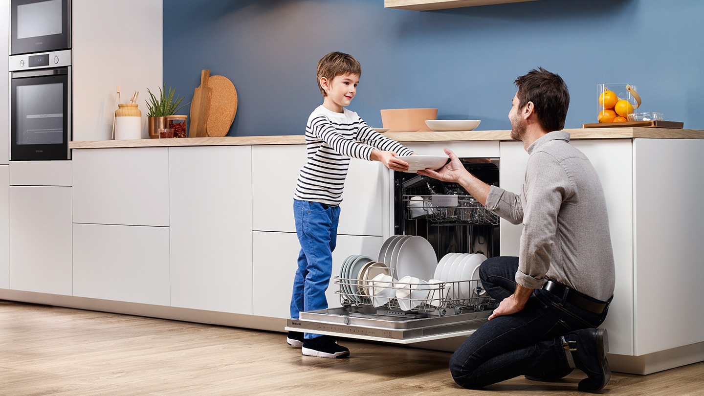 Un ragazzo sta passando dei piatti ad un uomo mentre la lavastoviglie in mezzo a loro è con lo sportello totalmente aperto.