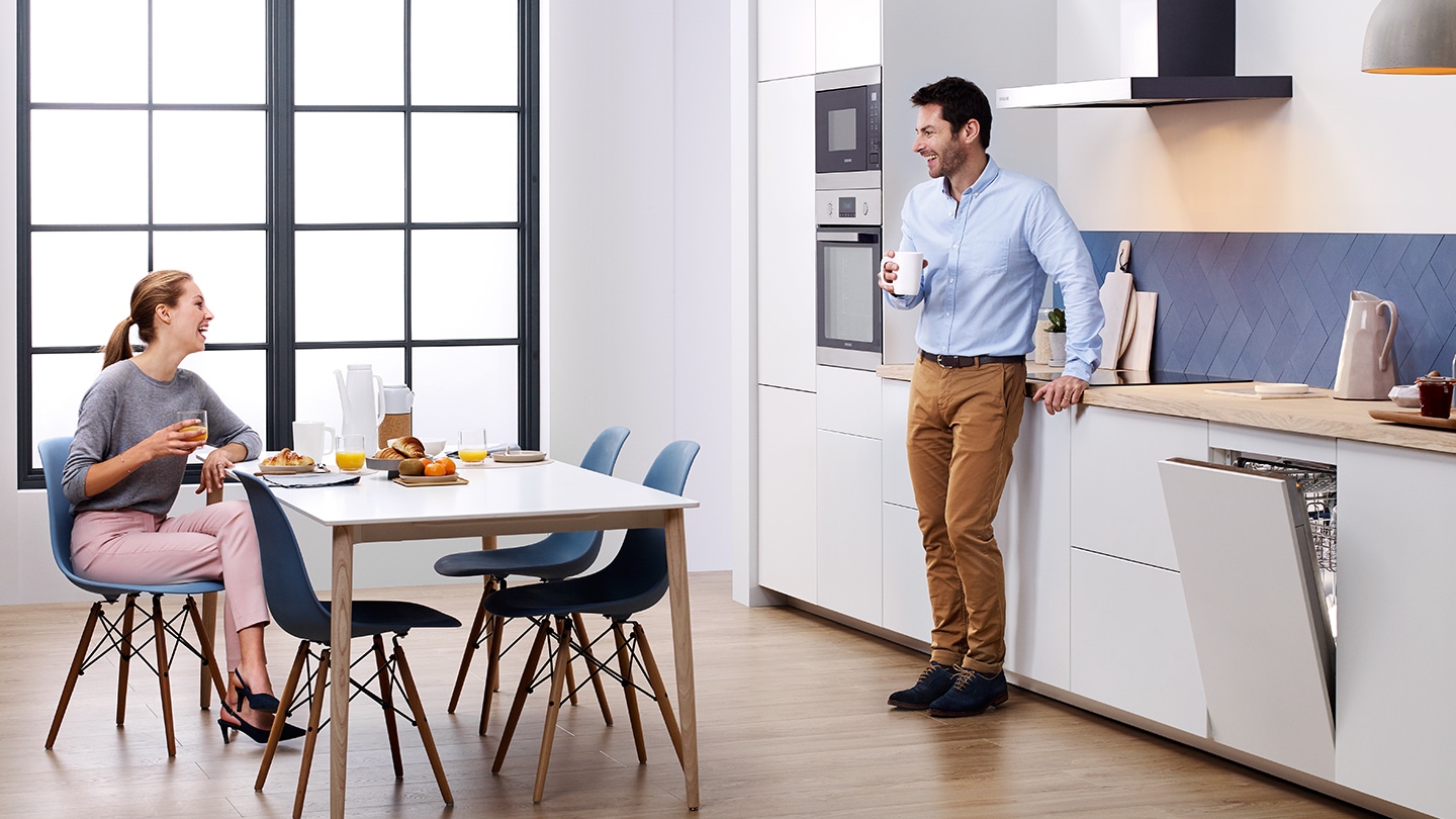 Una donna e un uomo stanno parlando all'interno di una cucina con gli elettrodomestici bianchi.