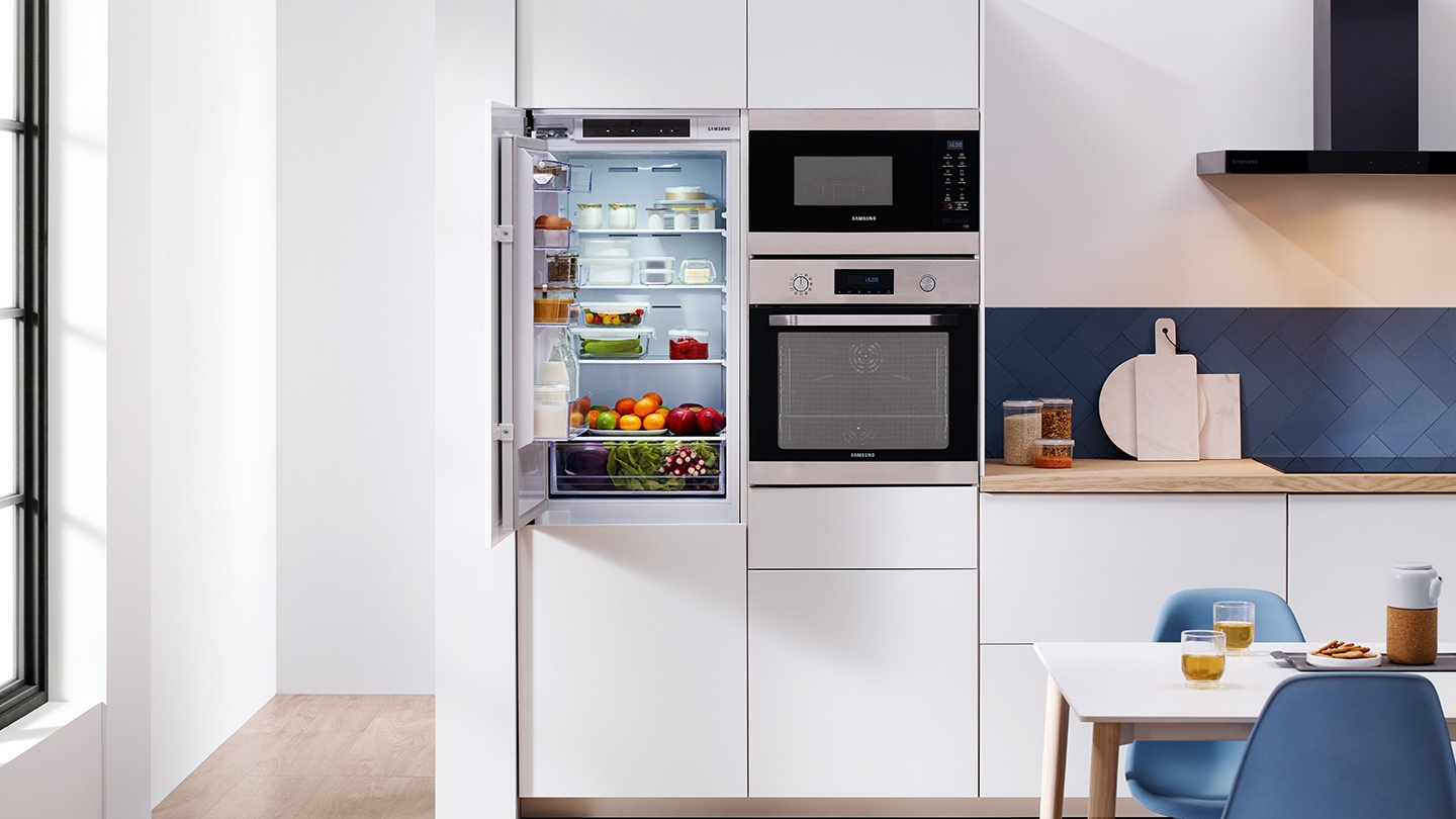 Un intero set di elettrodomestici da incasso Samsung come frigoriferi, forni, forni compatti,  installati in una cucina bianca premium mentre la porta del frigorifero è socchiusa.