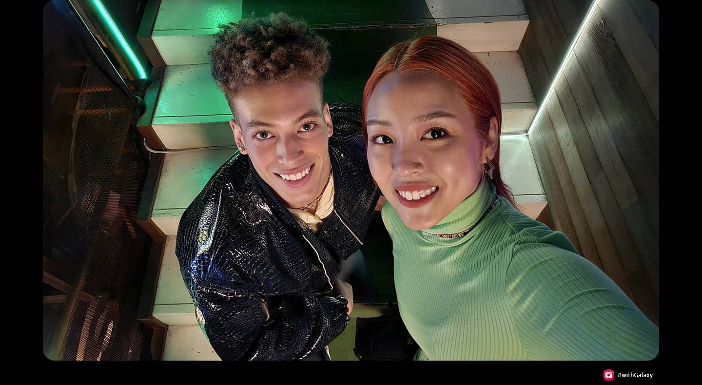 階段の上に並んで立ち、微笑みながら上を見上げている男女。夜の屋外で、照明は中央に立つ二人にフォーカスしている。女性は、写真を撮るためにカメラかスマートフォンを持つような格好で画面の外に腕を伸ばしている。下には、#withGalaxyのハッシュタグが表示されている。