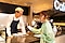外国のカフェで日本人女性と外国人の店員が会話している様子。女性の手にはGalaxy S24(ギャラクシーS24)のスマートフォンがもたれ、Galaxy AI(ギャラクシーエーアイ)の通訳機能を活用し、コミュニケーションを取っている。