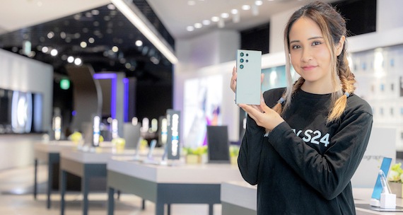 Galaxy Harajuku (ギャラクシー原宿)の店内で、Galaxy S24 Ultra(ギャラクシーS24ウルトラ)を両手で持ちながらこちらに微笑んでいる女性クルー。